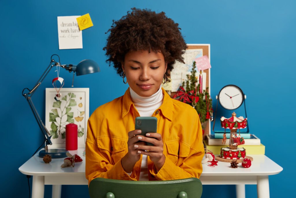 pleased-female-student-takes-break-from-autodidact-uses-cellular-chatting-online-browses-application-sends-text-message-checks-mail-via-wifi-sits-chair-near-workplace-blue-wall-2-1024x683.jpg